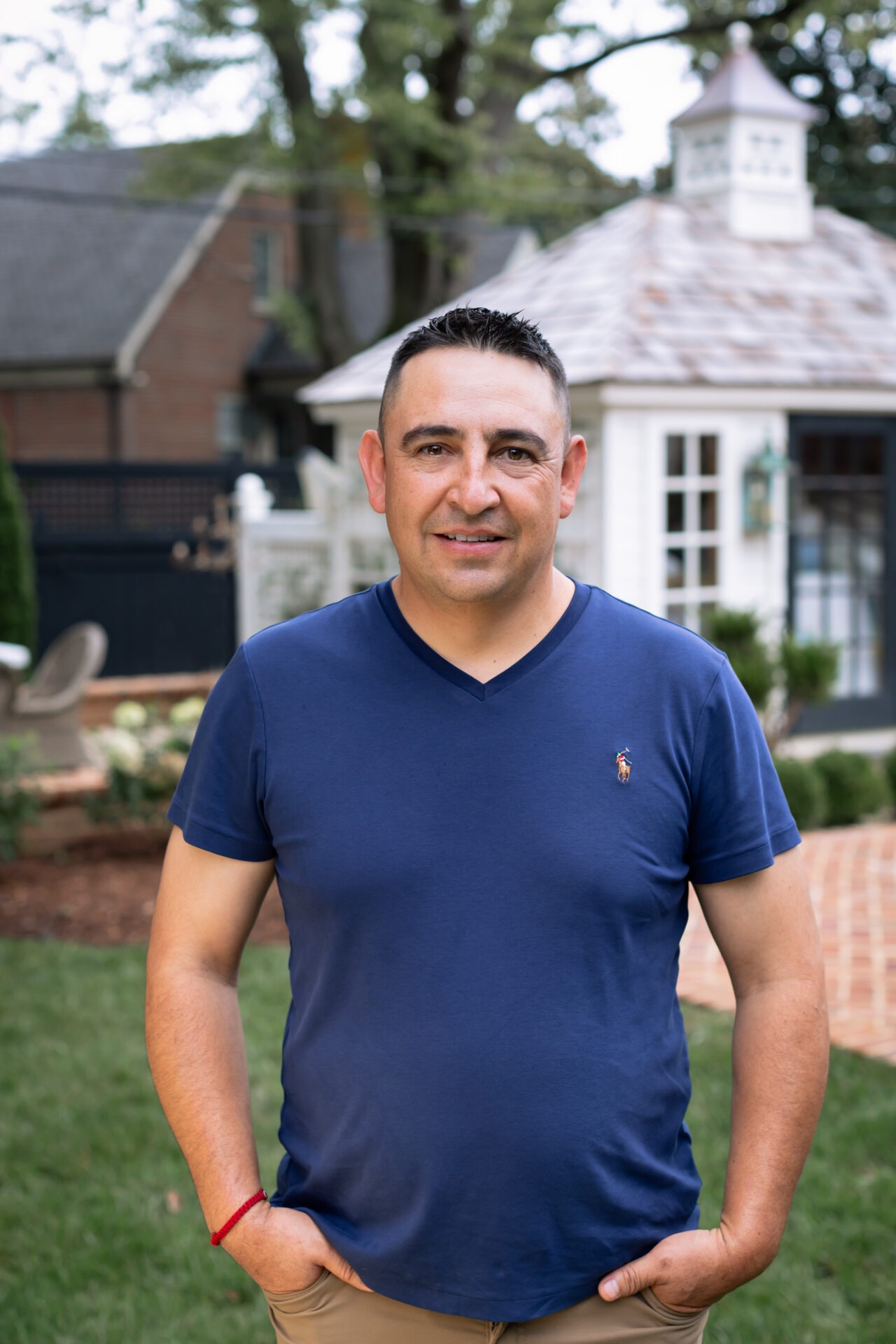 A person wearing a blue shirt stands in a garden with a small white building and greenery in the background.