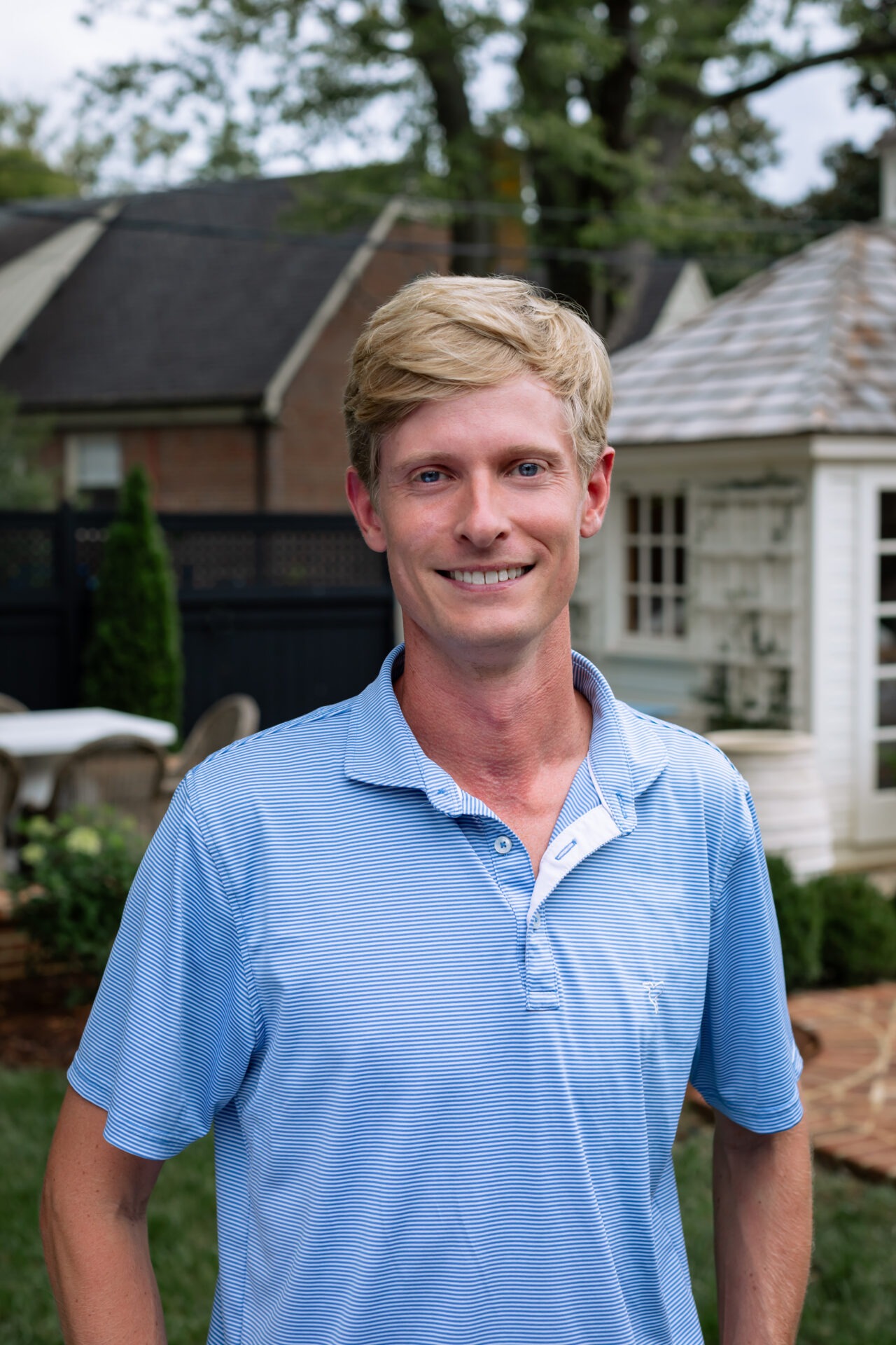 A person in a blue shirt stands smiling in a garden with patio furniture and a small building in the background.