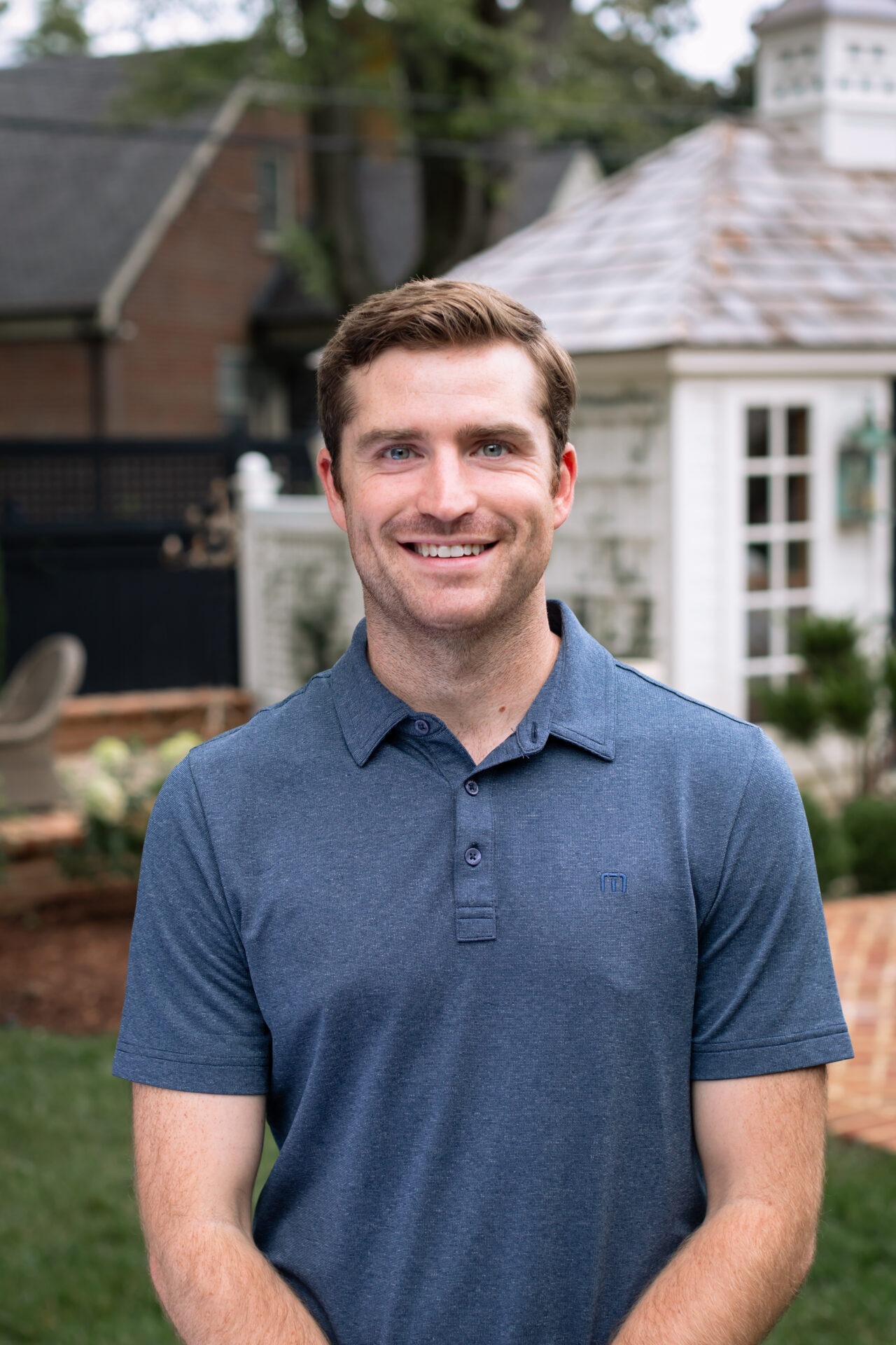 A person wearing a blue shirt stands in a garden with a white shed in the background, smiling at the camera.