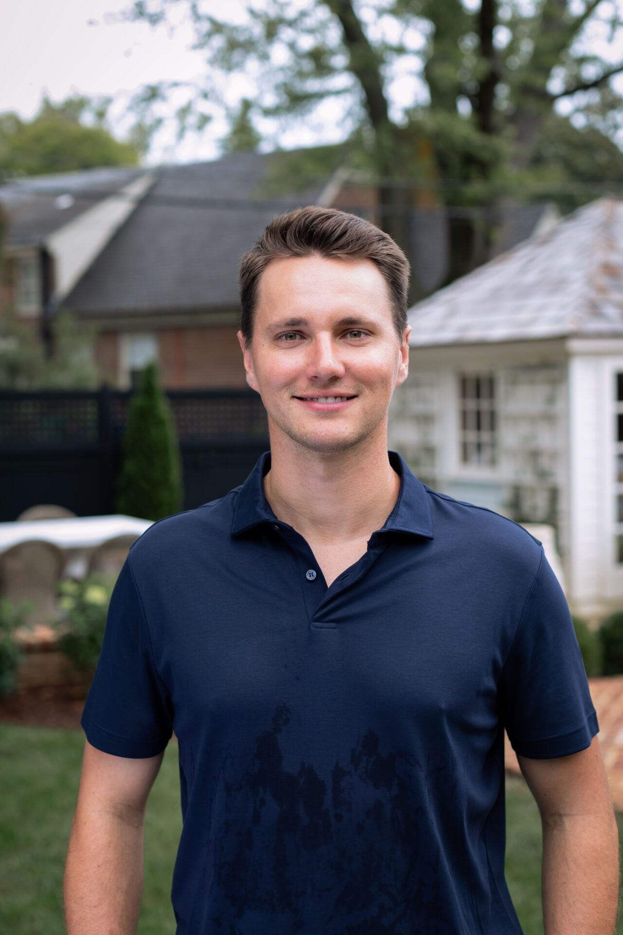 A person in a navy shirt stands outdoors in a residential backyard with trees and a small white building in the background.