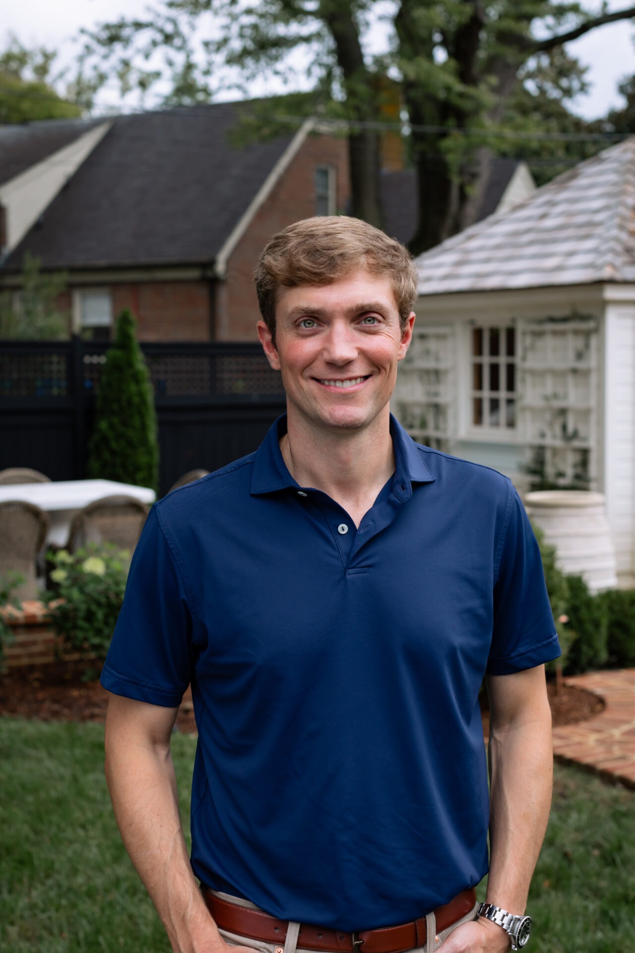 A person in a blue shirt smiles outdoors, standing in a garden with shrubbery, a small white building, and brick pathways nearby.