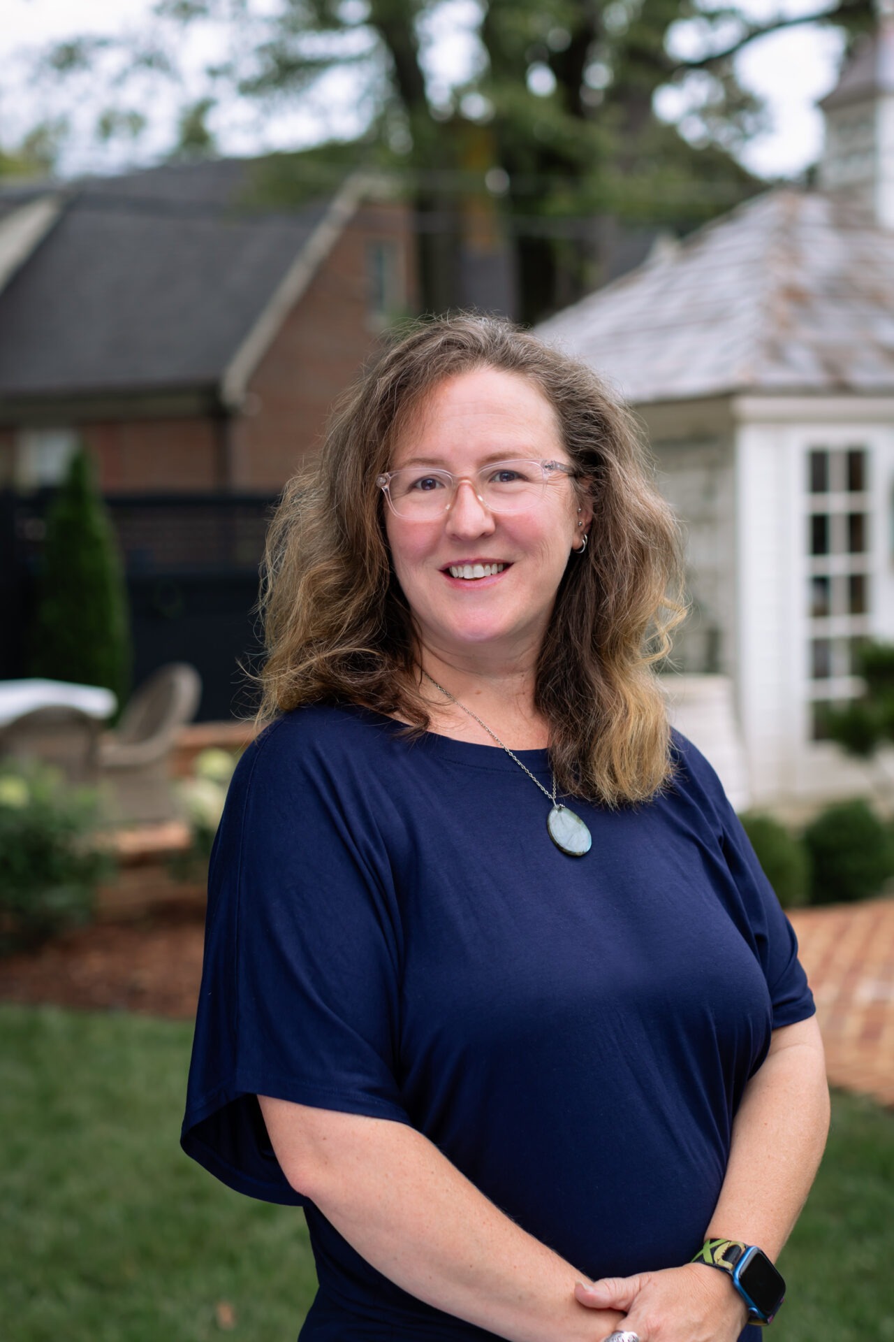 A person with glasses smiles in a garden setting, featuring a house with a peaked roof in the background.