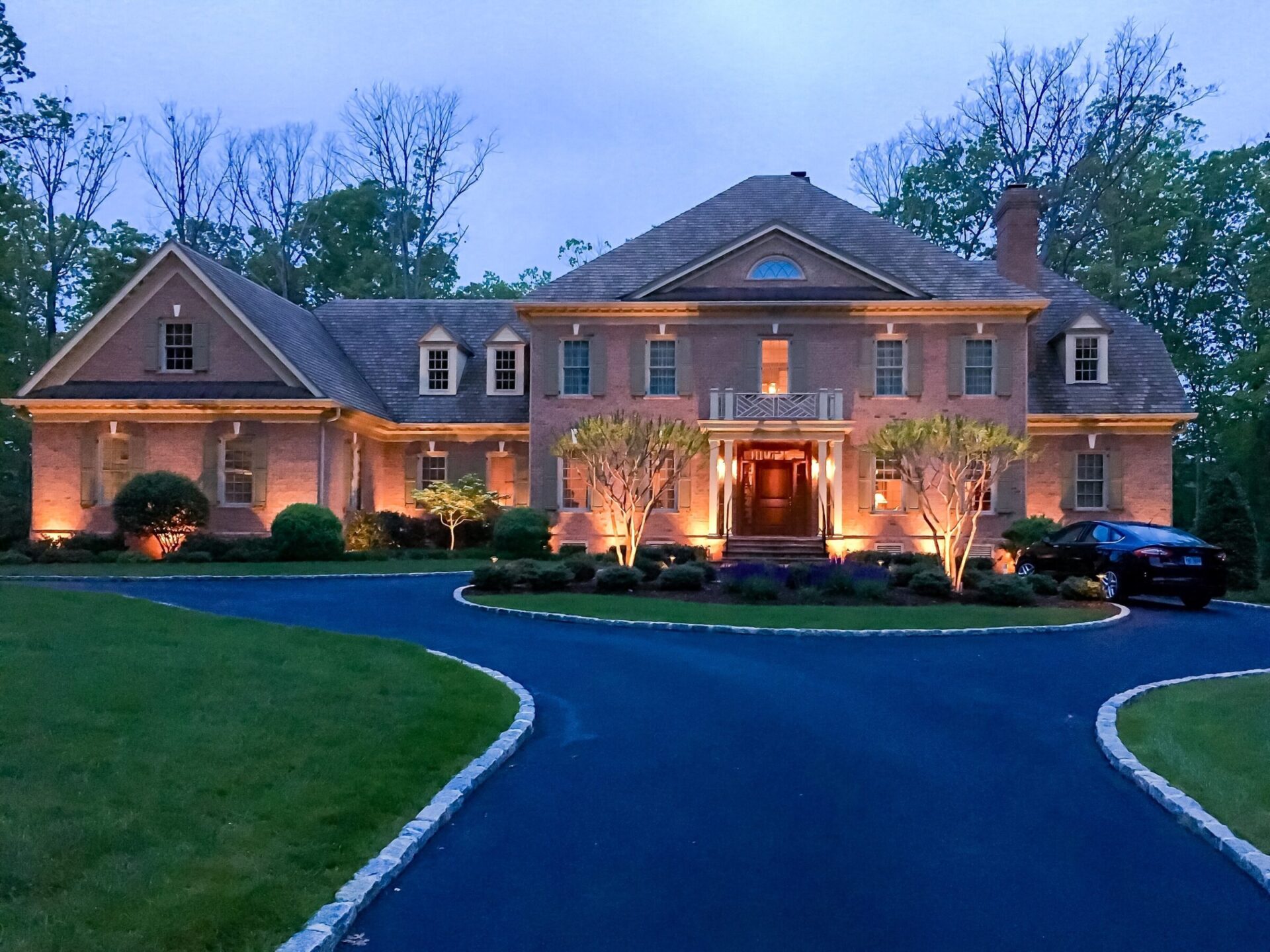 Elegant brick house with lit entrance and circular driveway, surrounded by trees and landscaped shrubs at dusk.