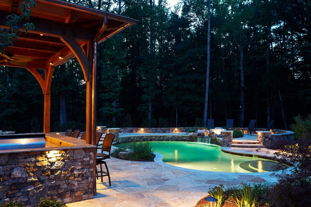 A serene evening scene with a stone patio, illuminated pool, and wooden pergola, surrounded by lush trees in a tranquil setting.