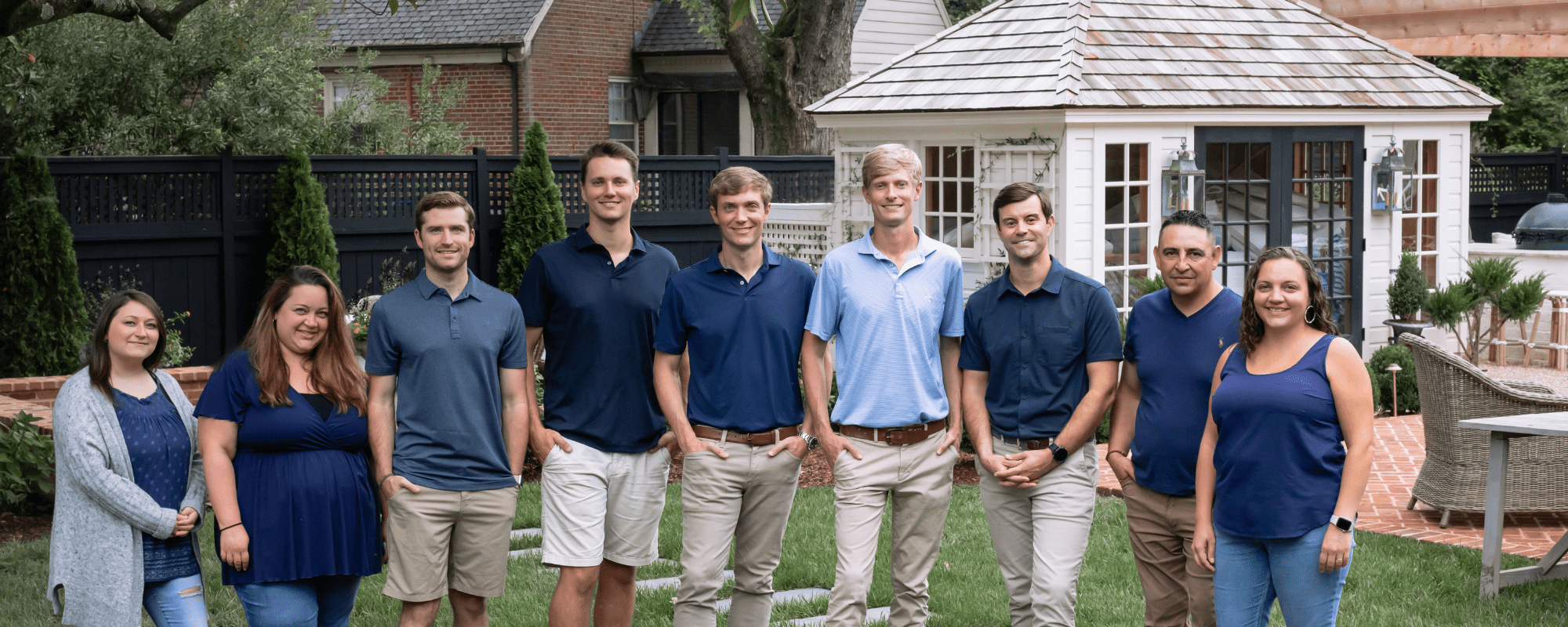 A group of eight people stands outdoors in front of a small white gazebo, surrounded by landscaped greenery and brick pathways.