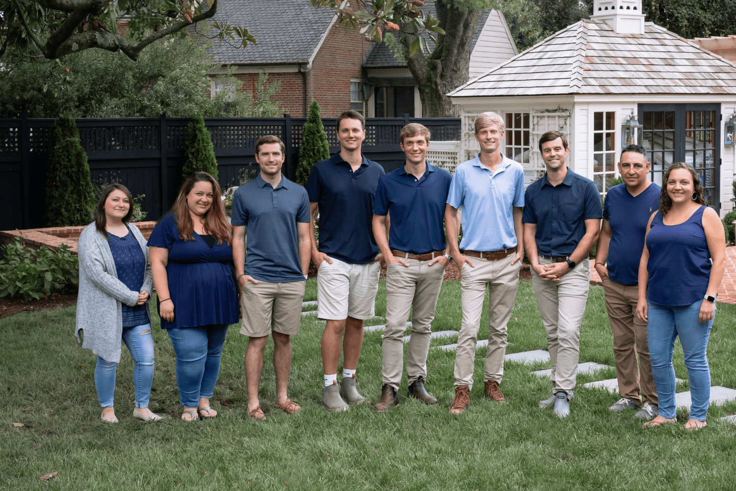 A group of nine people stand on a grassy lawn in front of a brick house and a white gazebo.