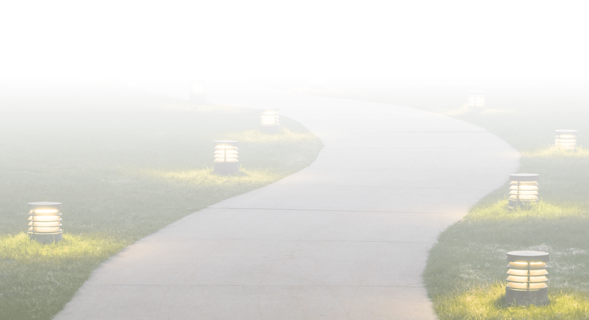 A foggy pathway with illuminated lanterns on both sides, winding through grass, disappearing into dense mist. No landmarks or people visible.