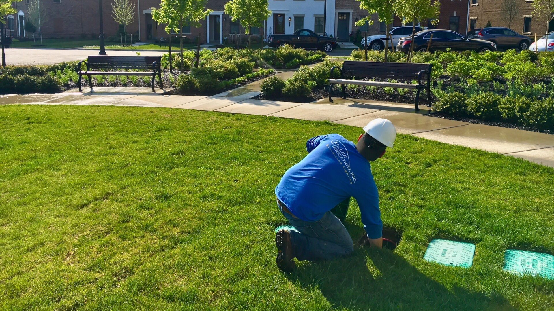 A person in a hard hat works on grass in a park with benches, trees, and parked cars in the background.