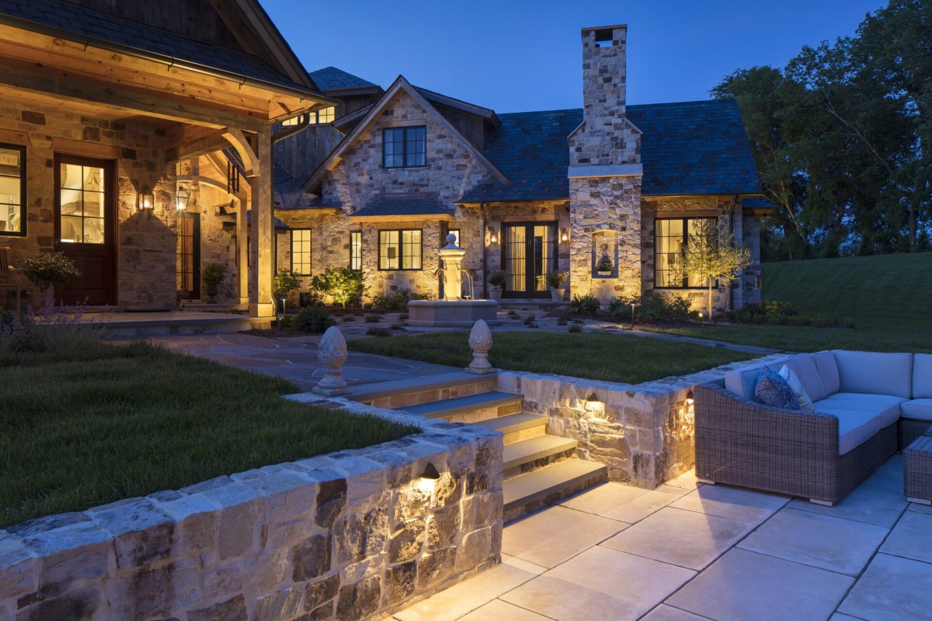 Charming stone house illuminated at dusk with cozy outdoor seating. Soft landscape lighting accents the architecture and lush greenery surrounding the home.