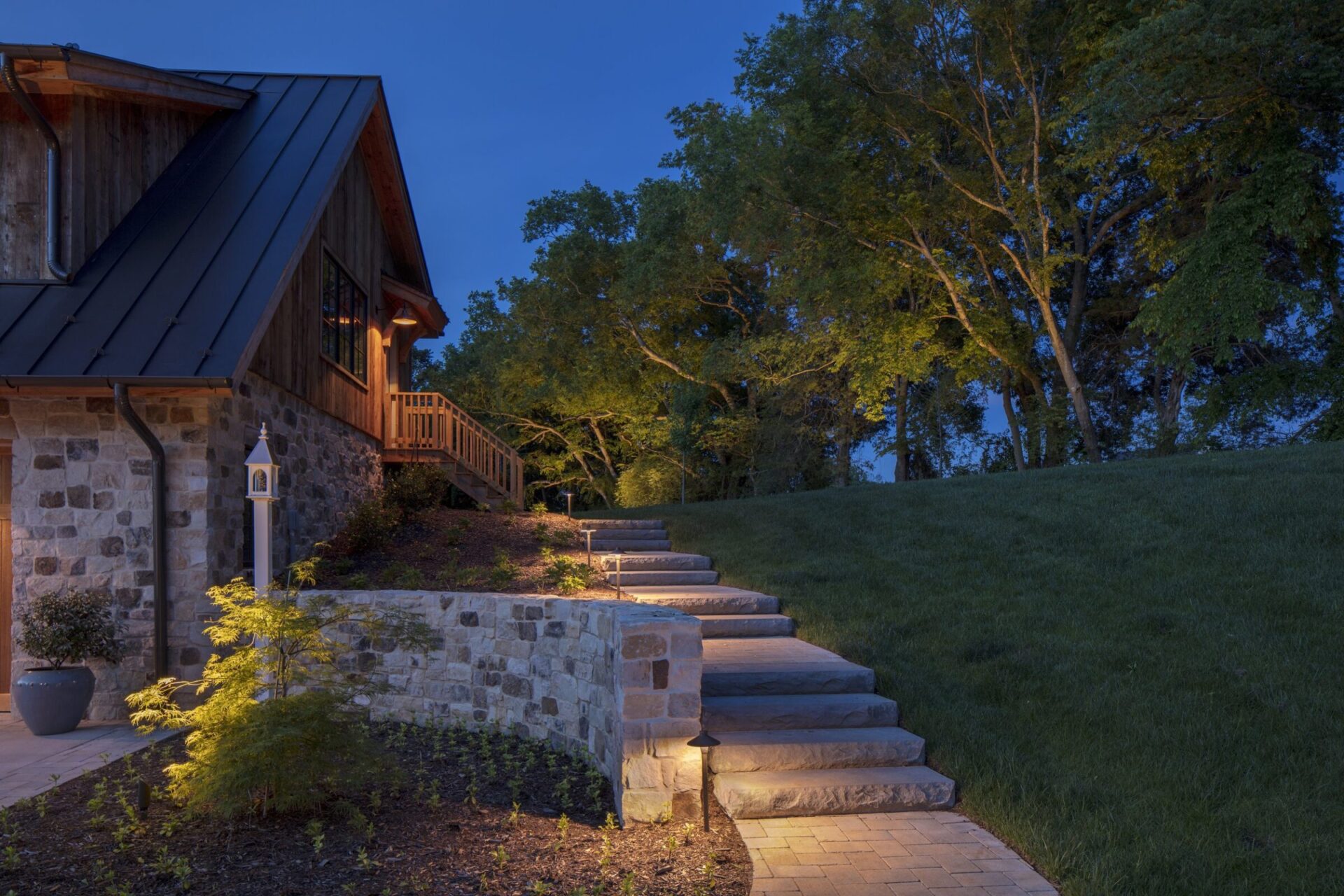 Charming stone house with wooden accents, set against a lush green landscape. Illuminated stone steps lead up to the entrance at dusk.