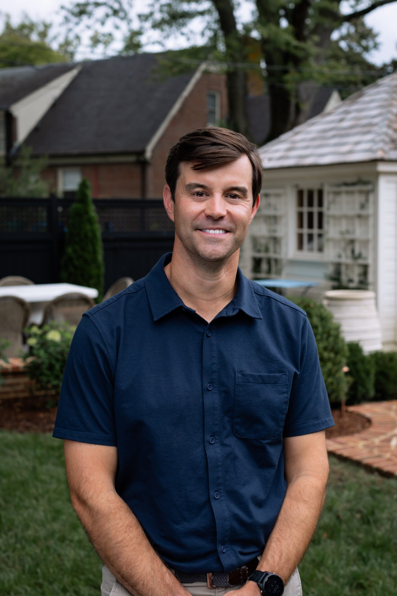 A person in a blue shirt stands in a garden with outdoor furniture, trees, and brick pathways, in front of a small building.