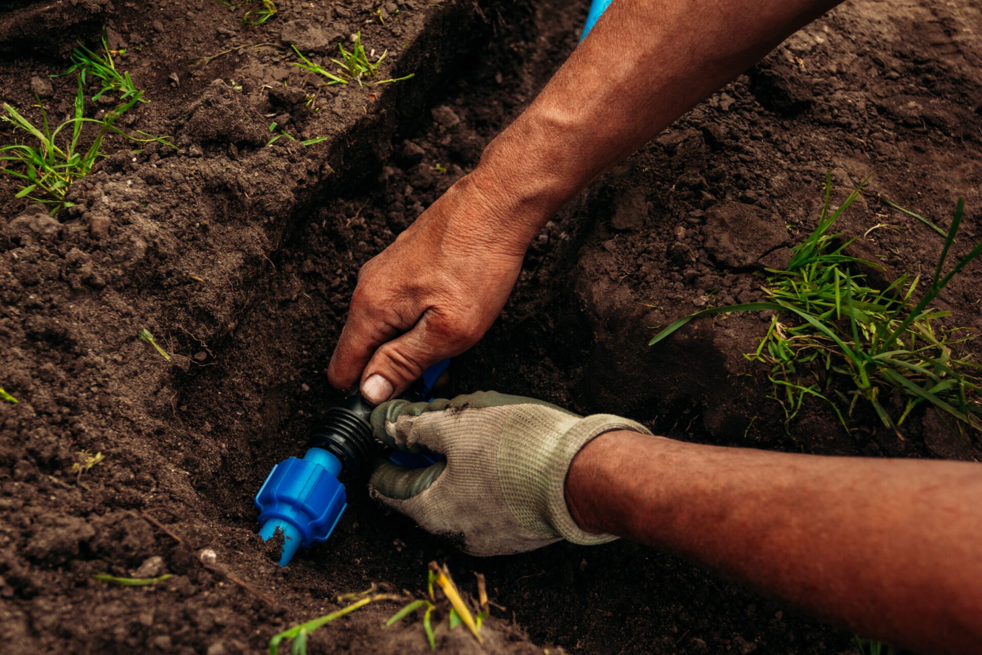 A person installs a blue irrigation connector in a trench surrounded by dirt and green grass, demonstrating agricultural or garden maintenance work.