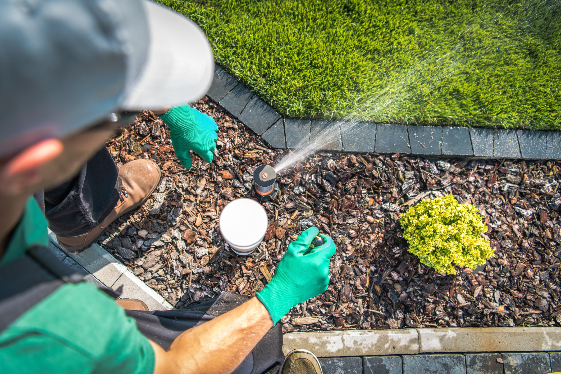 A person wearing gloves adjusts a garden sprinkler. There's neatly trimmed grass, a small plant, and a mulch-covered area in a landscaped yard.