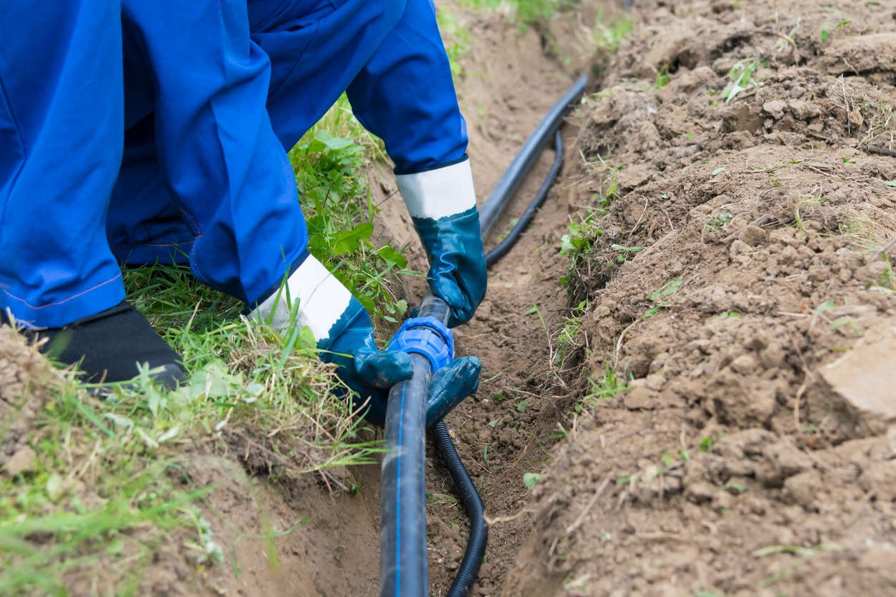 A person in blue overalls is connecting a black hose in a dirt trench, surrounded by grass and soil.