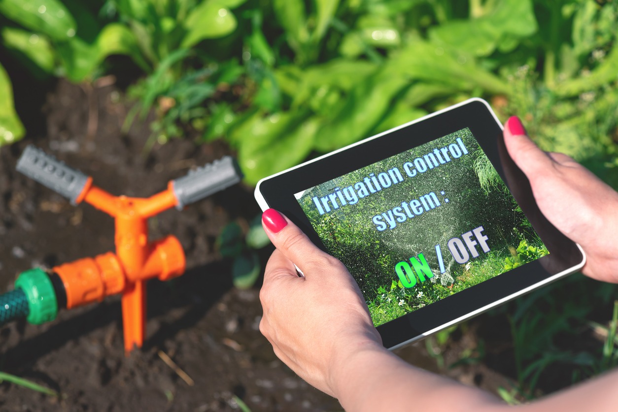 A person uses a tablet to control an orange garden irrigation system, surrounded by vibrant green plants. The tablet displays "Irrigation control system: ON/OFF."