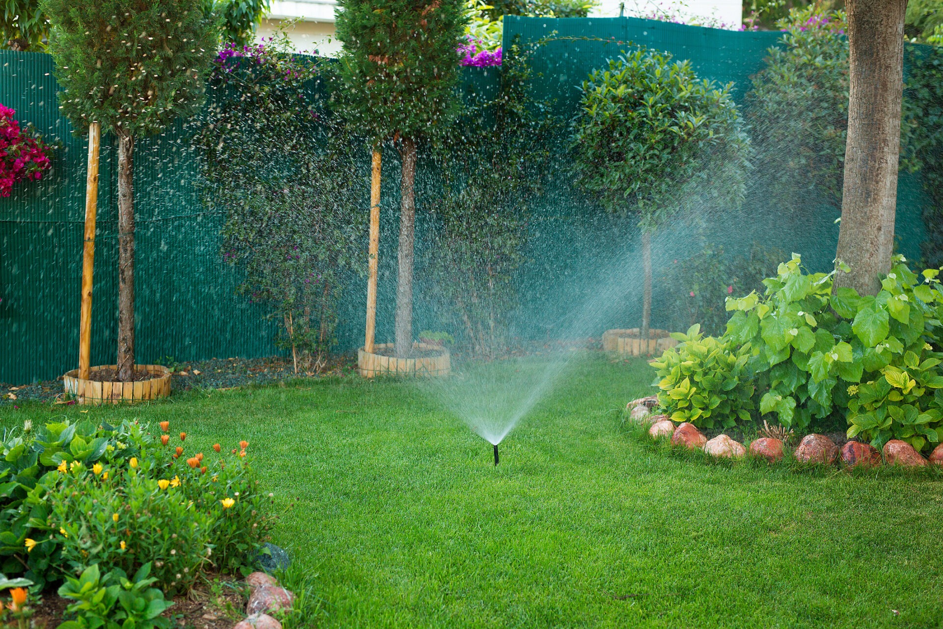 A lush garden with trees, vibrant flowers, and a sprinkler watering the green grass, surrounded by a fence and colorful bushes.