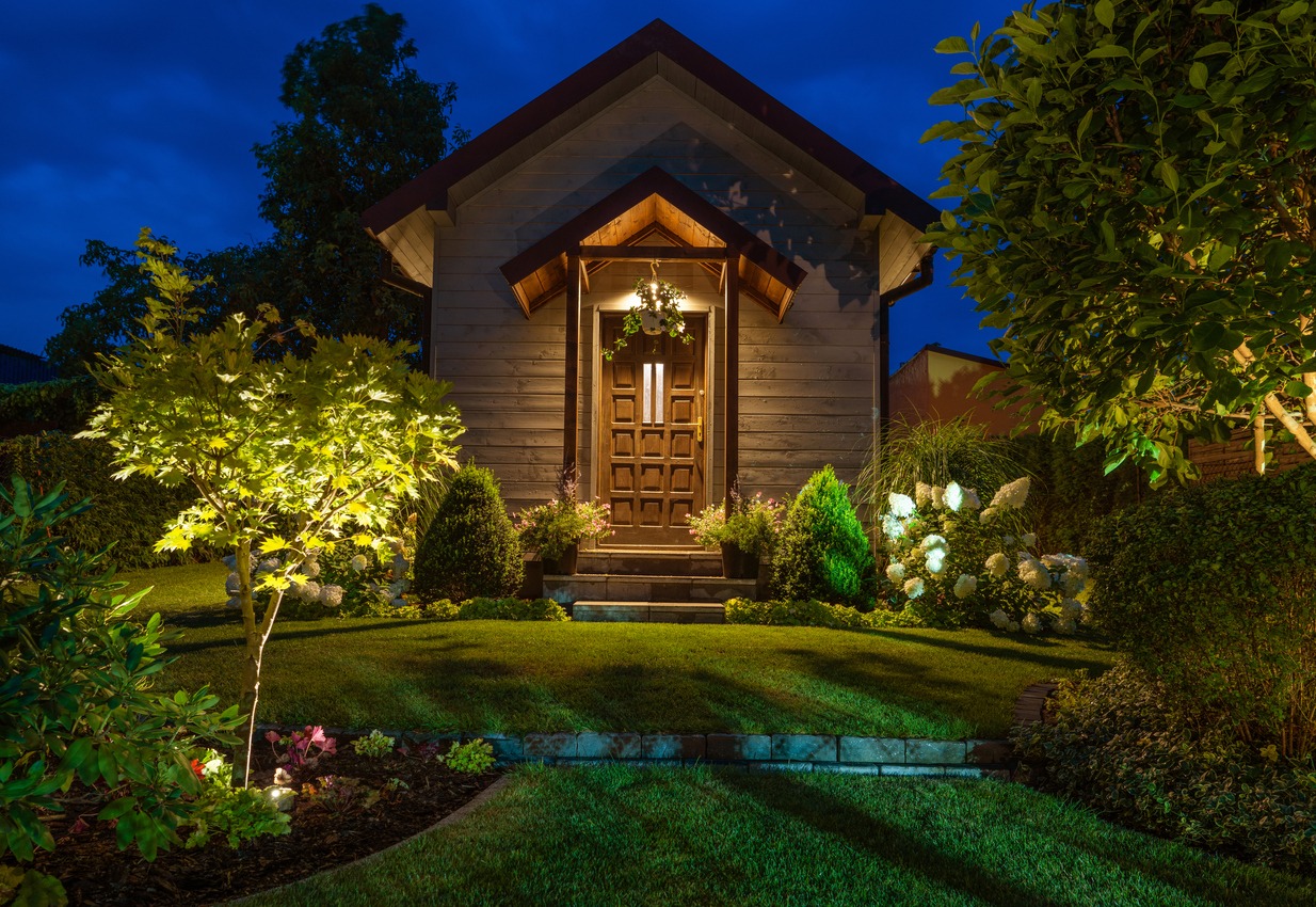 A small, wooden house with a warmly lit entrance is surrounded by a lush, meticulously landscaped garden under a clear evening sky.