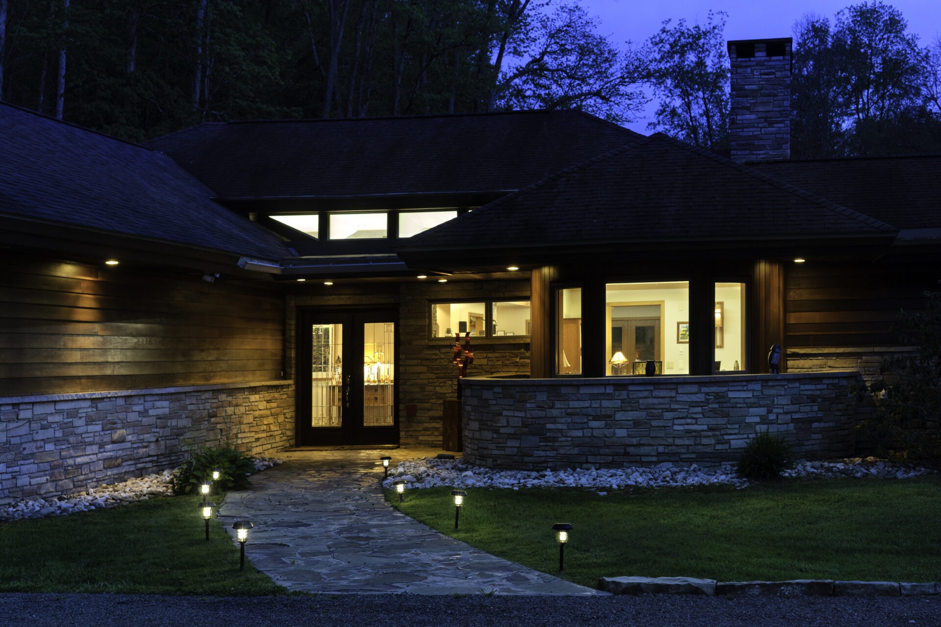 A modern house at twilight, with stone and wood exterior, glowing interior lights, surrounded by greenery and trees, creating a serene atmosphere.