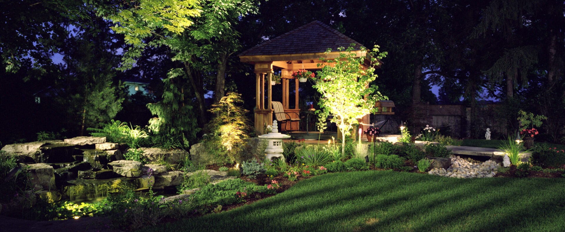 A beautifully lit garden at night features a wooden gazebo surrounded by lush greenery, stone pathways, and softly glowing landscape lights.