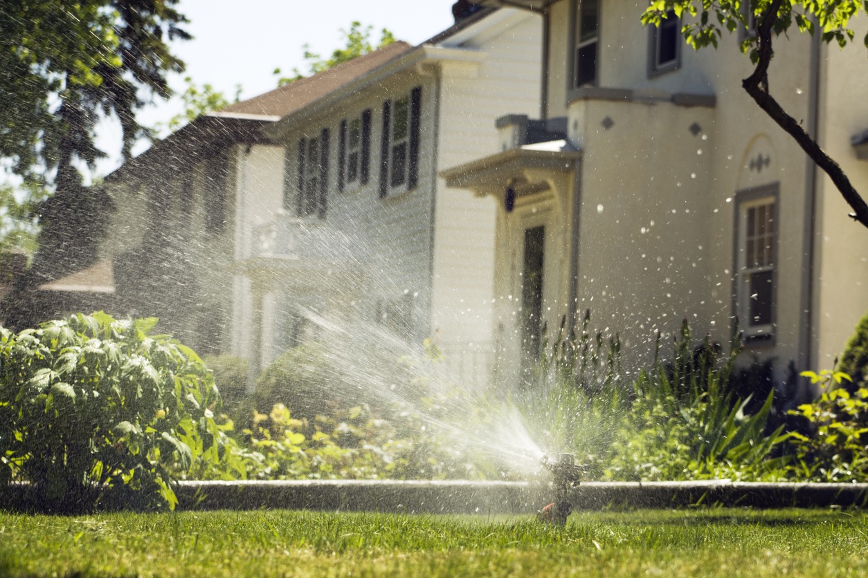 A suburban house with a lawn is being watered by a sprinkler, surrounded by greenery and sunshine, in a peaceful neighborhood.