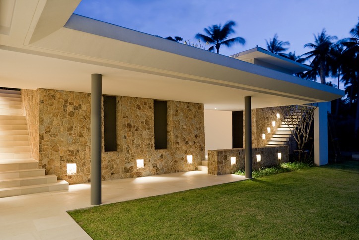 Modern house with stone walls and ambient lighting, featuring an outdoor staircase and surrounded by lush palm trees, under a twilight sky.
