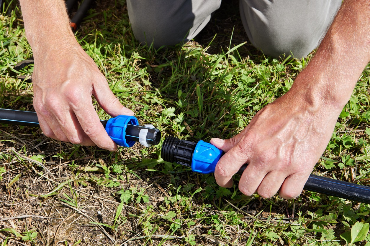 A person connects blue and black plastic hose fittings while kneeling on grass, focusing on irrigation setup or garden hose maintenance outdoors.