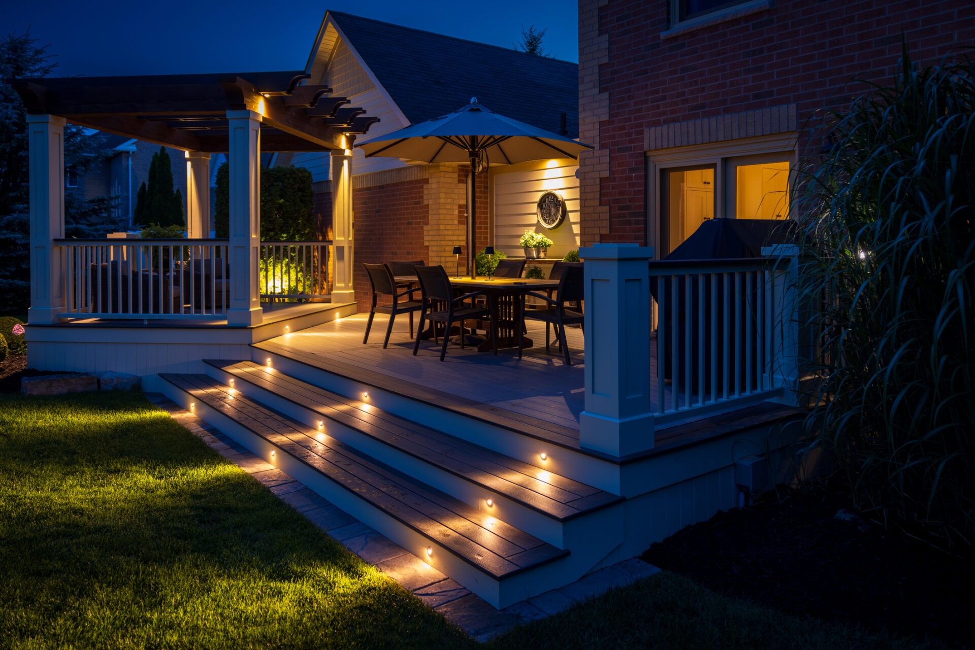 A well-lit patio at night features a dining table, chairs, umbrella, and a pergola. Soft lighting highlights the steps and garden.