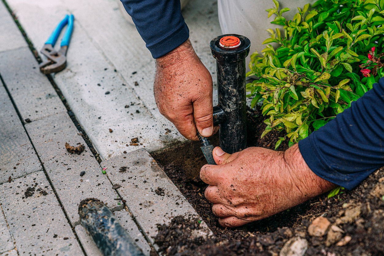 A person fixes a sprinkler system beside a walkway. Hands are visibly dirty, with tools nearby and greenery in the background.