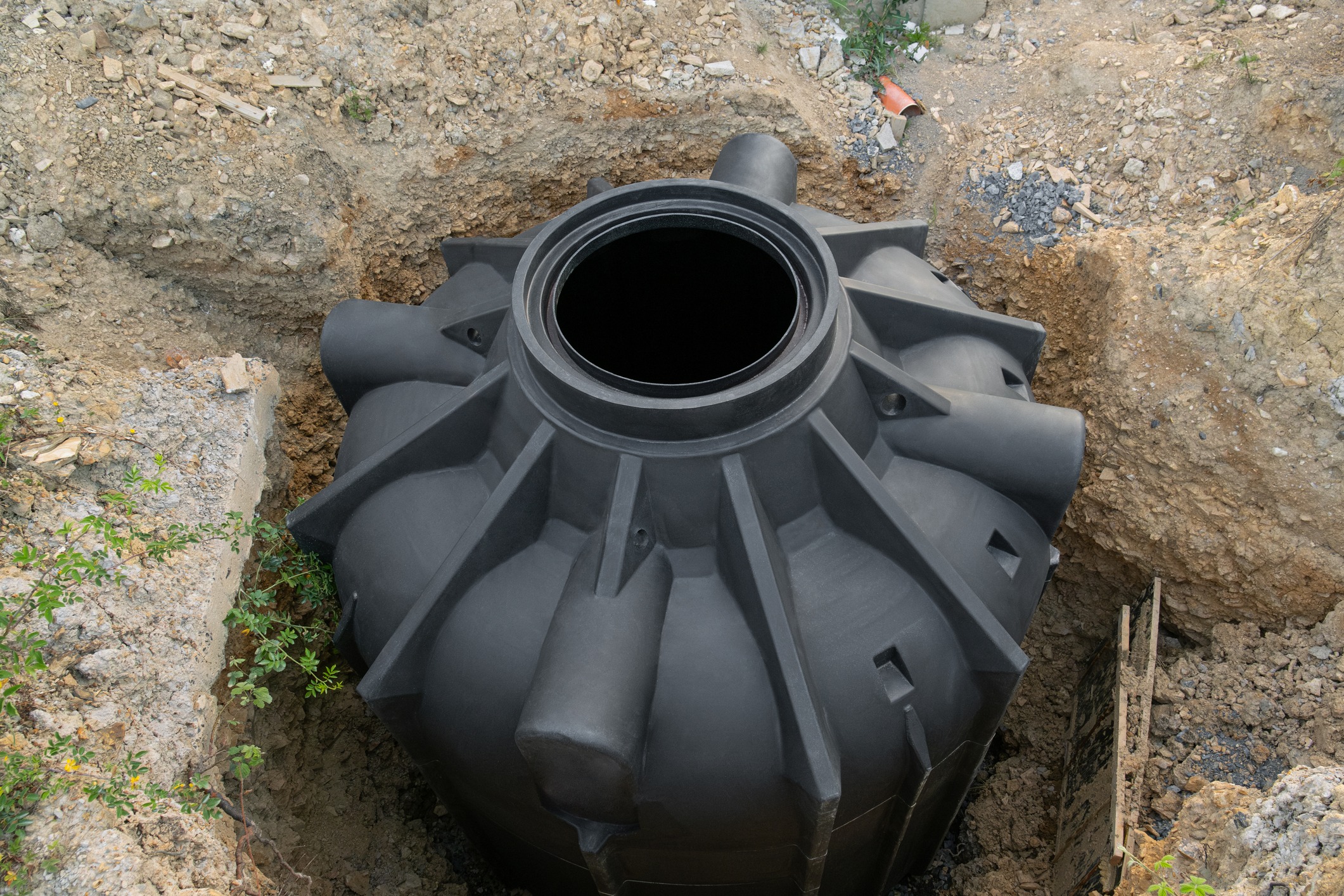 A large, black underground water tank is partially buried in the ground, surrounded by dirt and small plants, under construction.