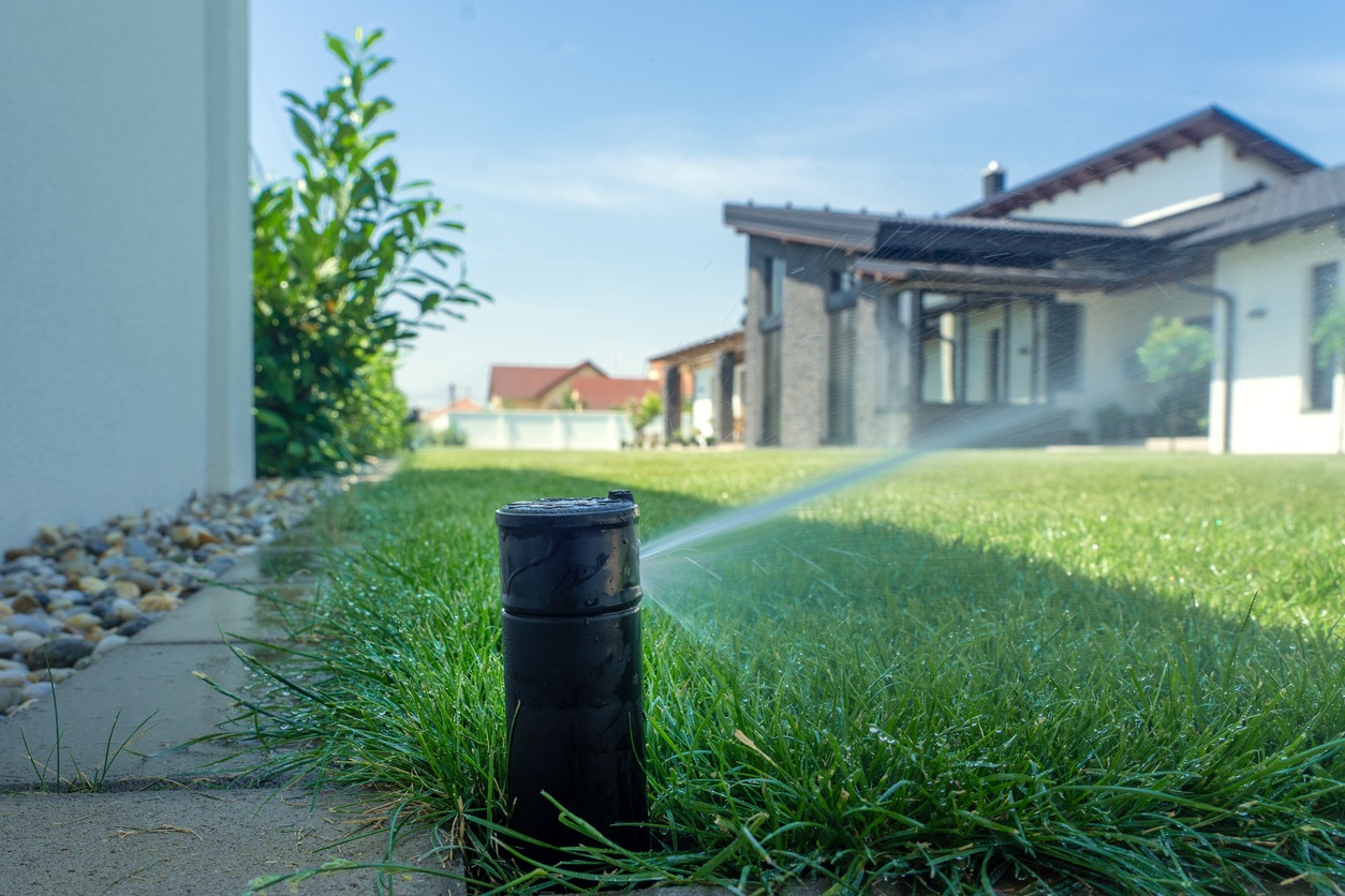 A lawn sprinkler waters a green yard in front of a modern house on a sunny day, with clear skies above.