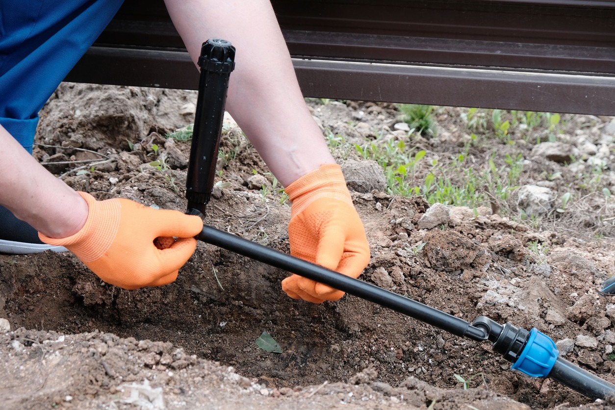 A person wearing orange gloves is installing or repairing a pipe in the soil, holding equipment for water or irrigation purposes.