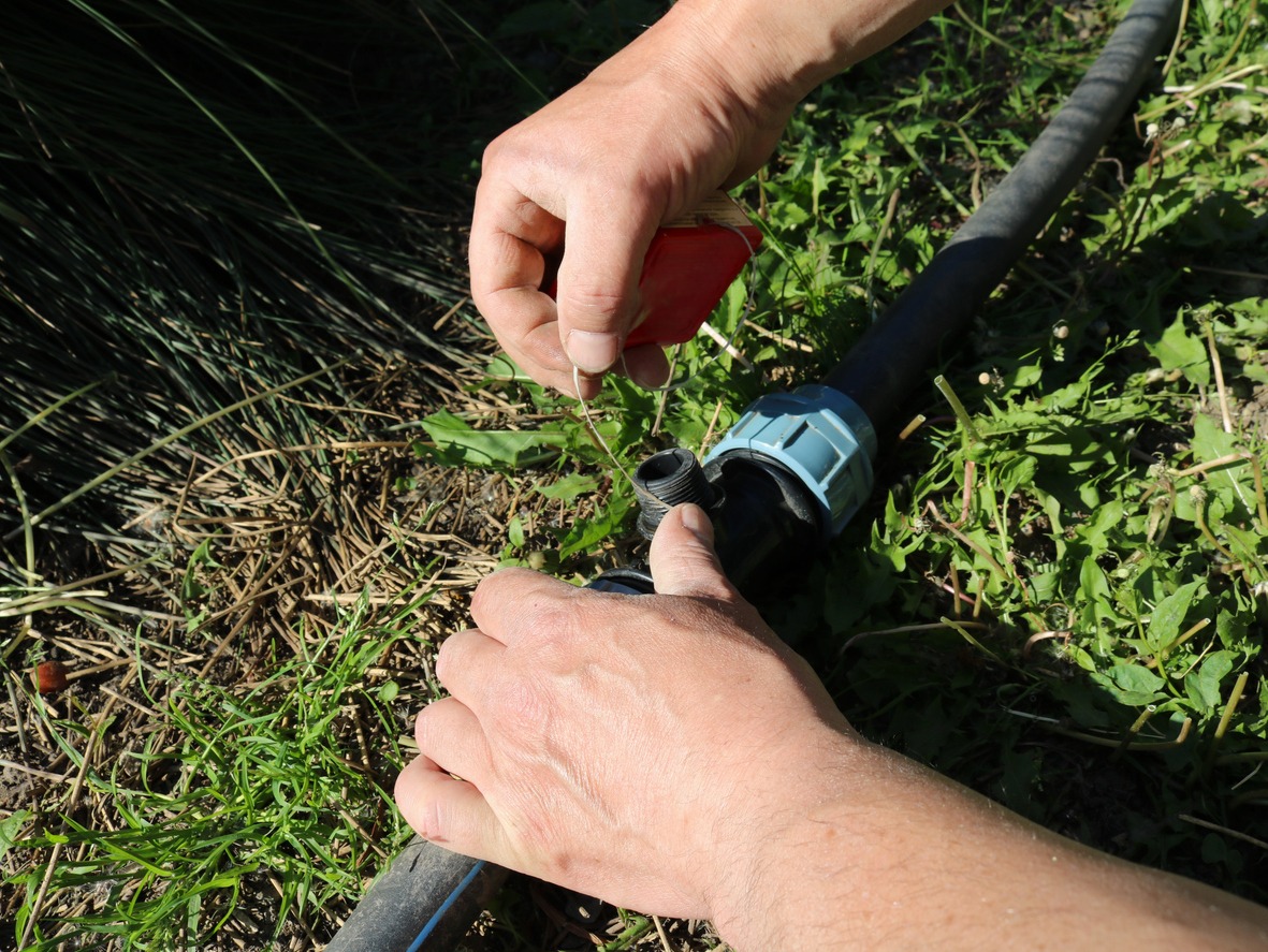 A person is repairing or inspecting a water hose connection outdoors, surrounded by grass. Hands are using a tool on the hose.