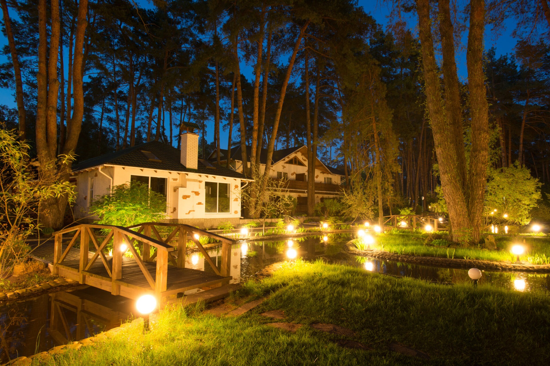 A serene nighttime garden scene features illuminated pathways, small wooden bridges, and cozy houses nestled among tall trees, creating a peaceful, enchanting atmosphere.