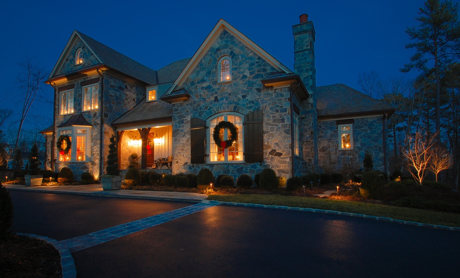 Stone house with lit windows and festive wreaths, surrounded by trees at dusk. Soft outdoor lighting accentuates driveway and landscaping. Quiet, elegant ambiance.