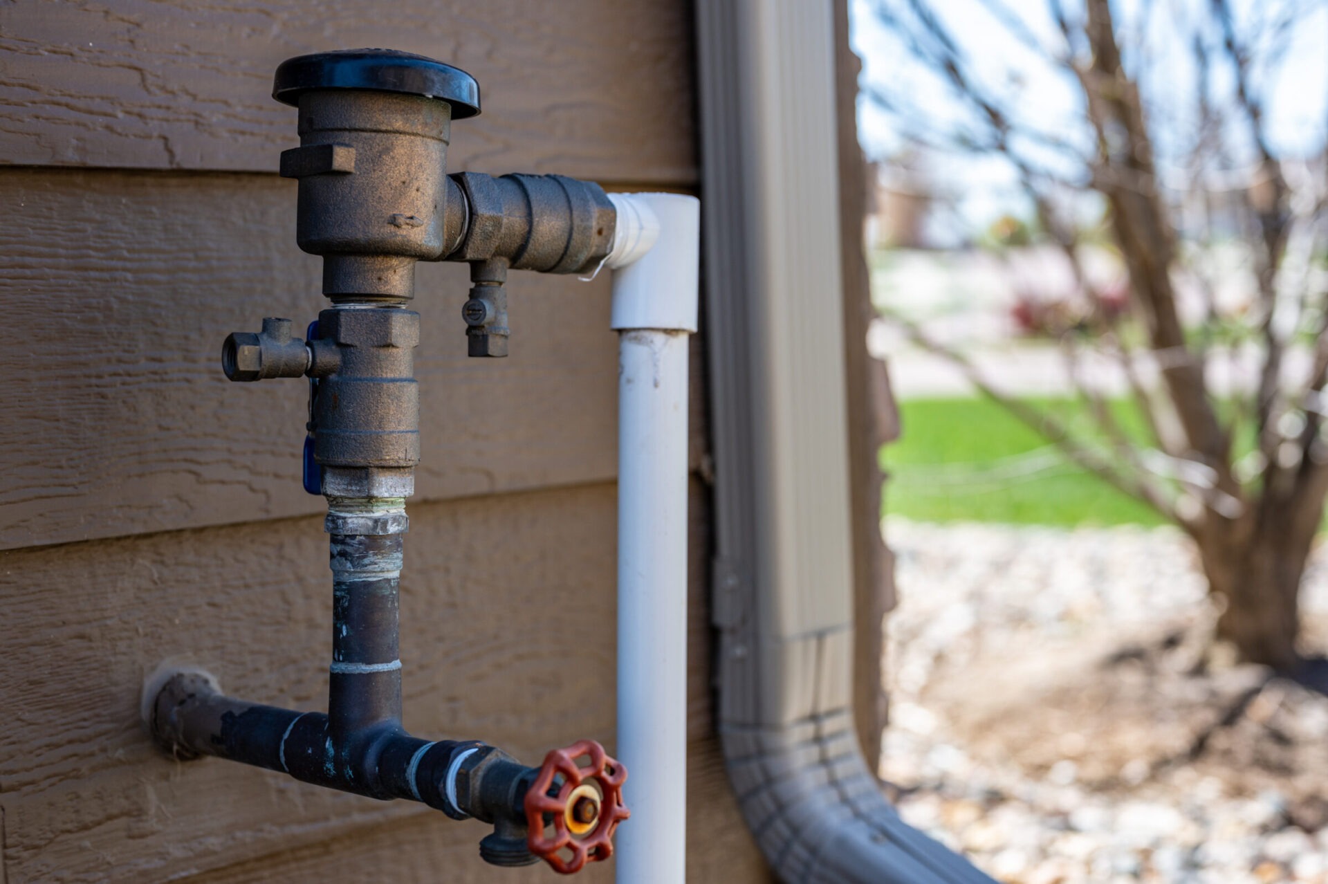 An outdoor water spigot with a rusty valve attached to a house exterior. In the background, there is a bare tree and grassy area.