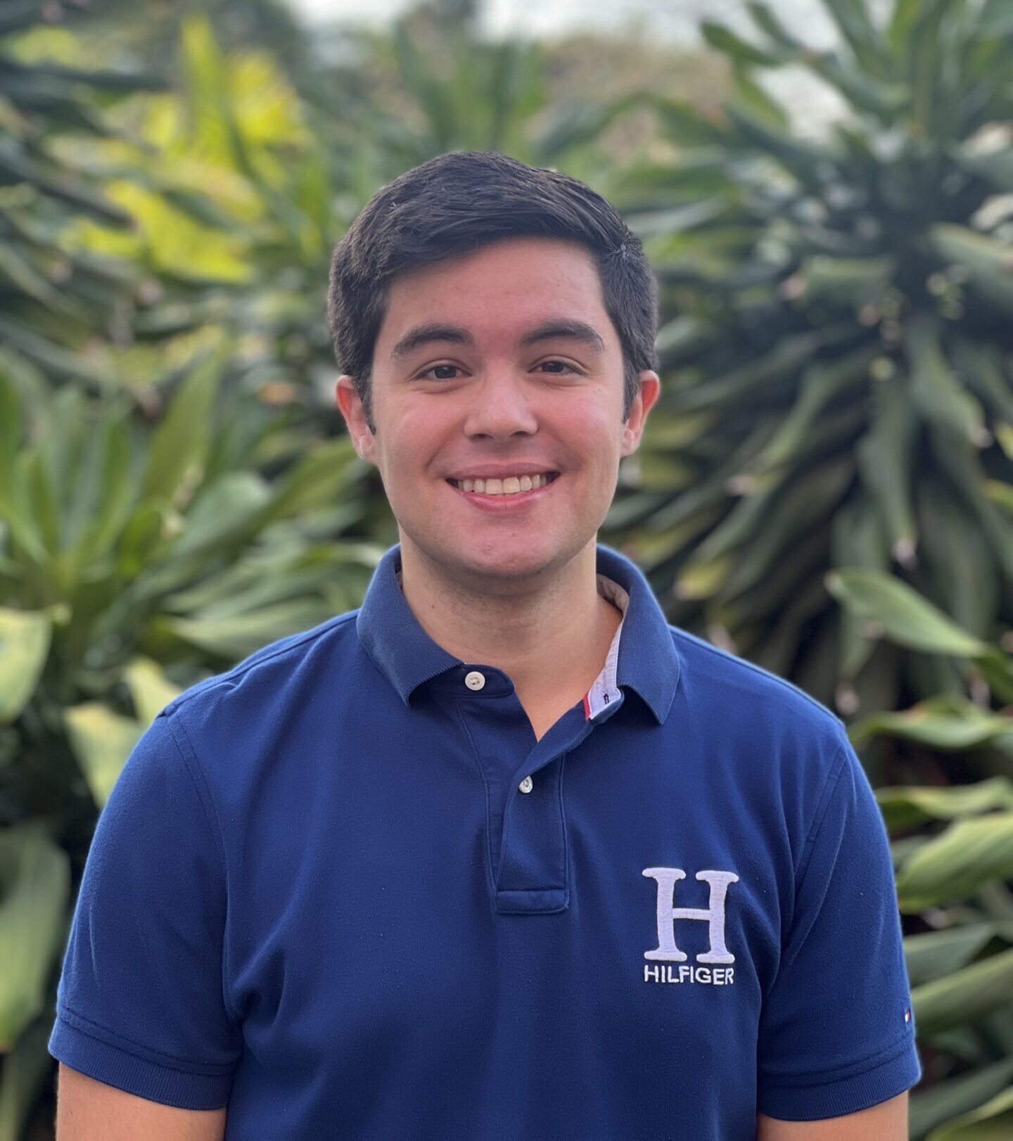 A person in a blue shirt stands smiling in front of lush green foliage, with no recognizable landmarks or historical buildings visible.