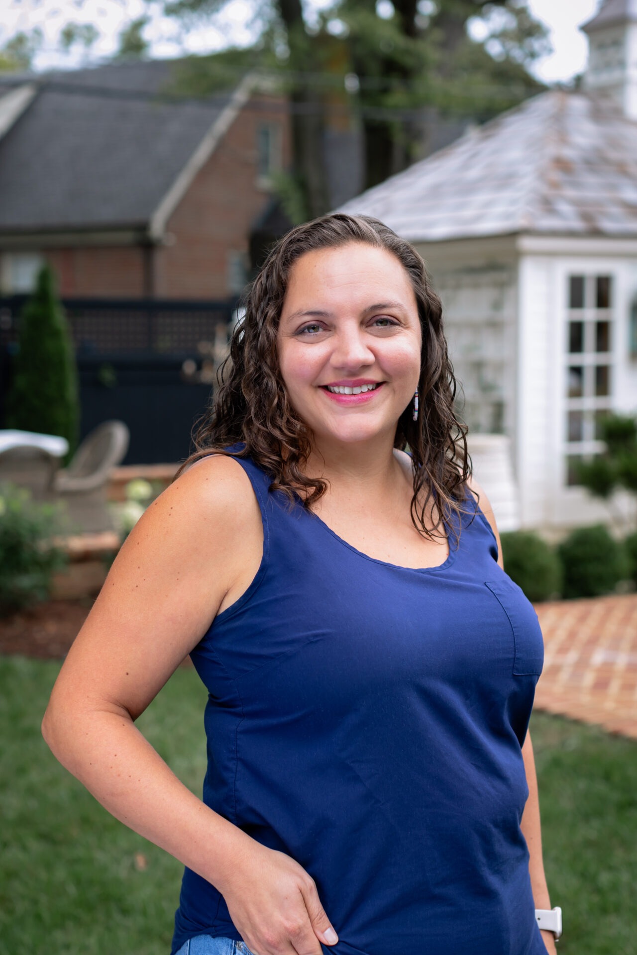 A person in a blue shirt stands smiling in a garden with a small white building in the background.