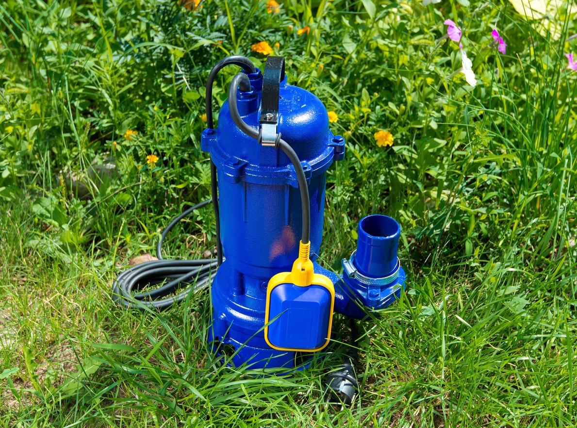 A blue submersible pump with a attached float sits on green grass surrounded by blooming yellow and pink flowers under sunlight.