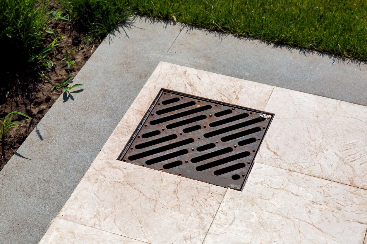 A metal drain cover set in marble tiles, surrounded by a grassy area. Sunlight highlights the textured surface and vibrant green grass.