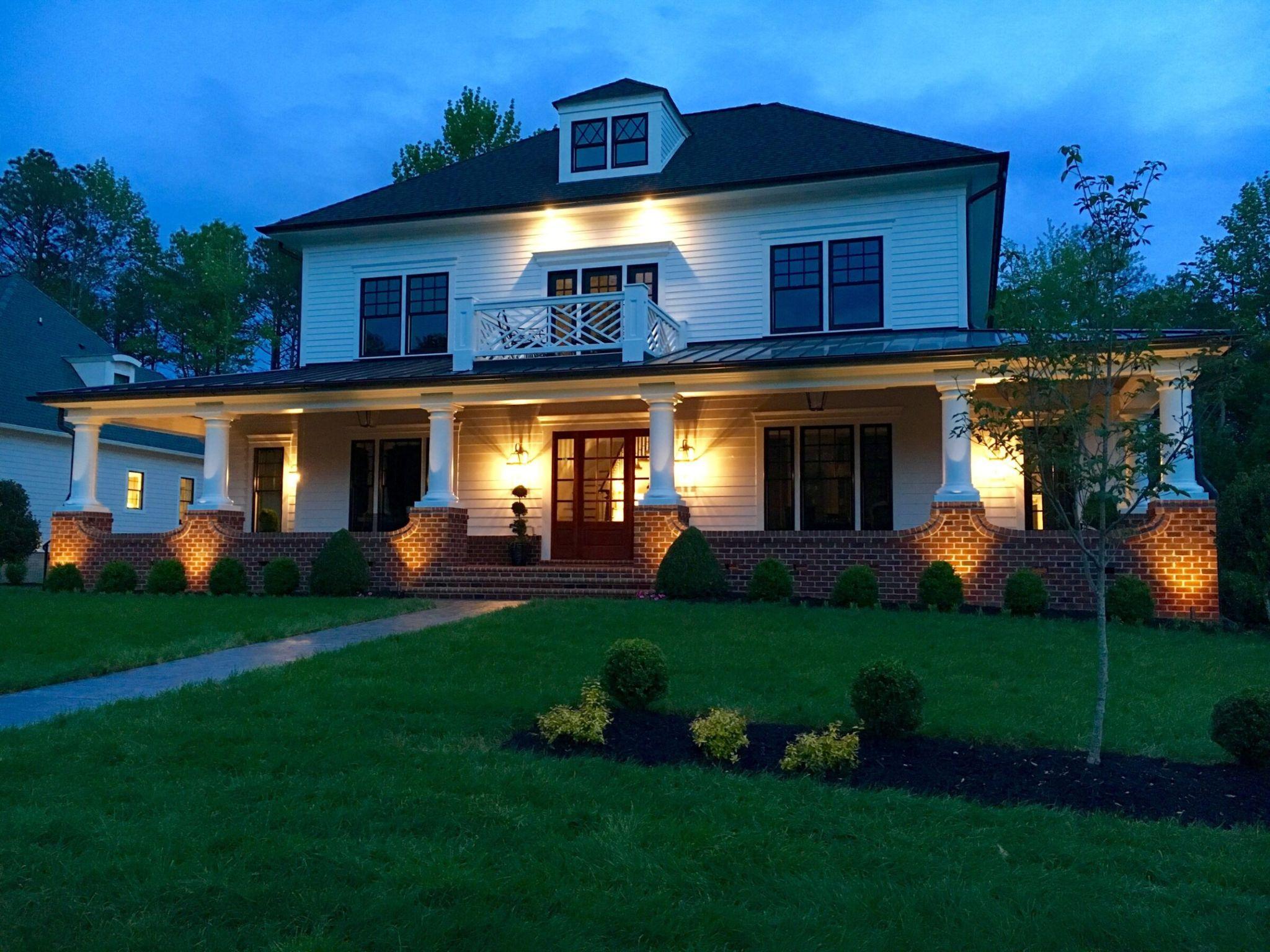 A two-story house with porch lights on, surrounded by a landscaped yard and trees, under a dusky blue evening sky.
