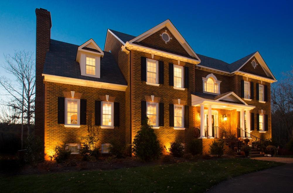 brick home exterior with warm landscape lighting illuminating the front facade, entry columns, and surrounding shrubs at dusk