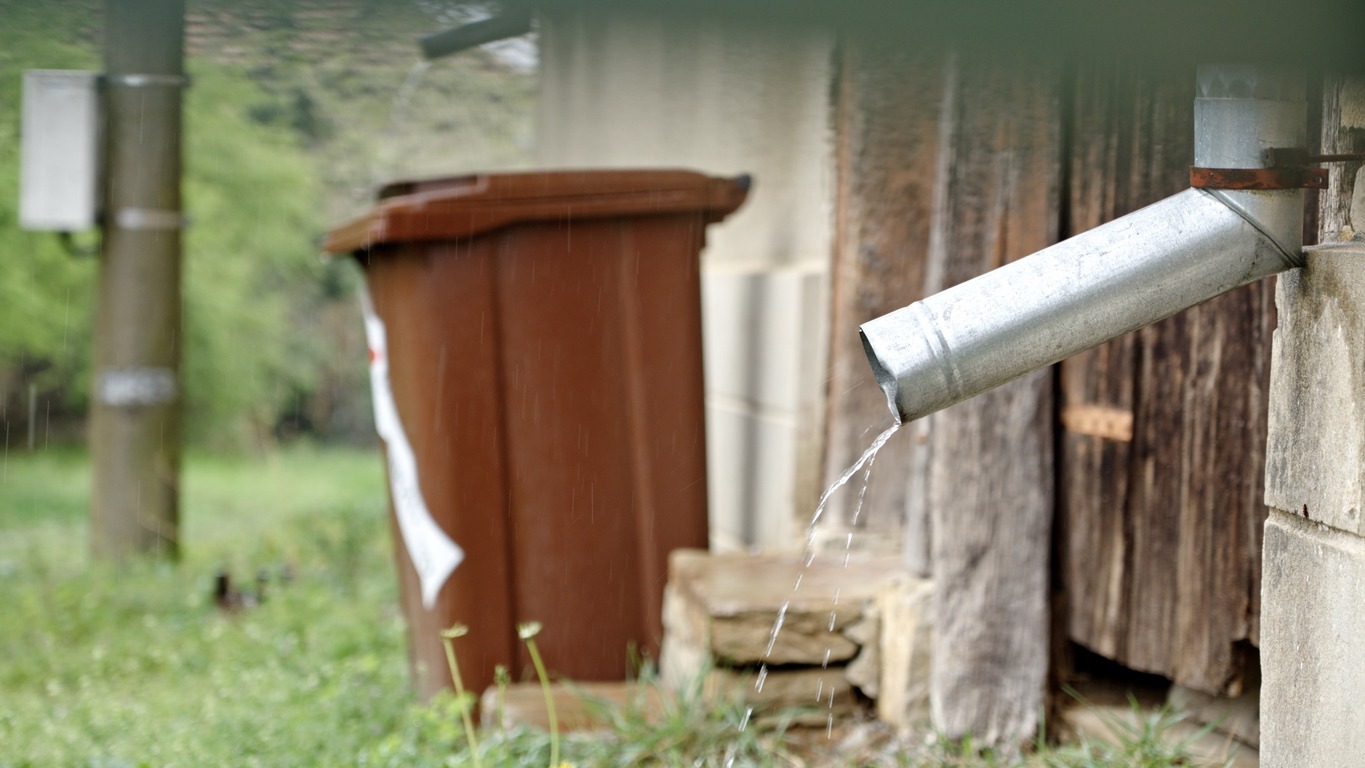 A metal gutter drains water beside a brown trash bin. The setting is outdoors, next to a wooden and brick structure, surrounded by grass.